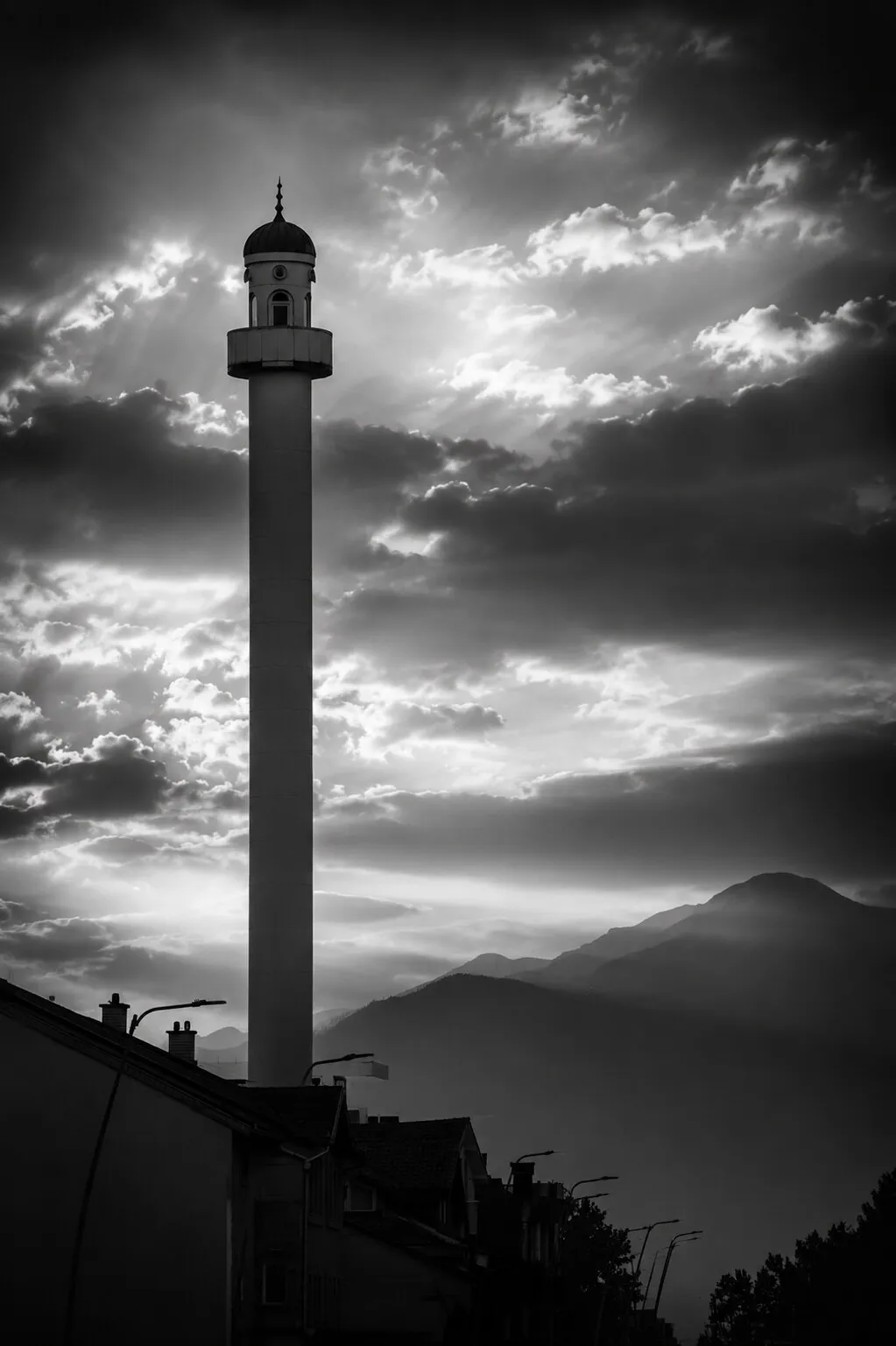 A tall, slender mosque minaret rising against a dramatic evening sky, mountains behind.
